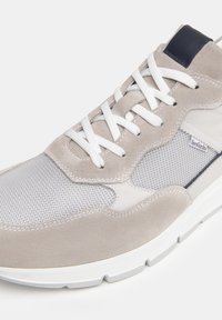 Zapatilla deportiva de color beige claro con materiales de ante y malla, cordones blancos, cuello acolchado y un acento en contraste de color marino en el talón.