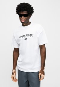 Λευκό βαμβακερό μπλουζάκι με την επιγραφή «new balance ATHLETICS» σε μαύρο. Το μοντέλο φοράει μαύρα γυαλιά ηλίου, έχει υφή δέρματος και γκρι παντελόνια.