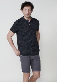 Crna majica s kratkim rukavima, polo stil, s geometrijskim uzorkom u plavim linijama. Uparena s svijetlosivim šortsevima, oboje napravljeno od lagane tkanine.
