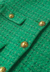 Primo piano di un tessuto verde texturato con bottoni dorati; presenta un motivo intrecciato e una superficie leggermente rialzata per una maggiore dimensione.