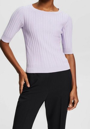 Kobieta ubrana w jasnofioletowy prążkowany sweter z krótkim rękawem i czarne spodnie, z jedną ręką opartą na biodrze, a drugą u boku.