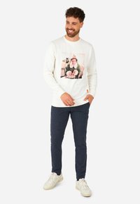 Vit sweatshirt med en stor grafisk tryck av en leende man i en grön tomteklädnad, ihopparad med mörka texturerade byxor och vita sneakers.