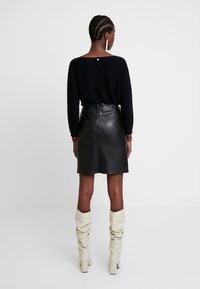 Žena s copy v černém svetr, černé kožené sukni a krémových vysokých botách stojící na jednobarevném pozadí.