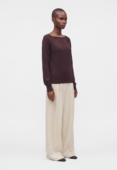 Personne debout portant un haut à manches longues foncé et un pantalon large beige avec des chaussures noires sur un fond clair uni.