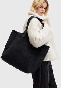 Sac fourre-tout en polaire noire avec une texture douce, doté d'une grande ouverture et de poignées en sangle noire. Logo subtil embossé sur un côté.