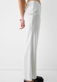 Dlouhá, těsně přiléhající bílá sukně z hladké látky, s bočním rozparkem a čistým siluetou, doplněná černými botami.