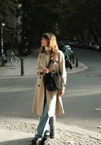 Beiger Trenchcoat mit schwarzen Knöpfen, getragen über einem weißen Rollkragenpullover und blauen Jeans. Das Modell hält eine schwarze Bucket Bag und steht auf einer Straße.