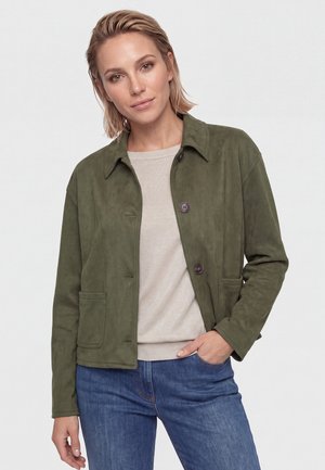 Frau mit kurzen blonden Haaren trägt eine olivgrüne, geknöpfte Jacke über einem beigen Oberteil und blauen Jeans, steht mit einer Hand in der Tasche.
