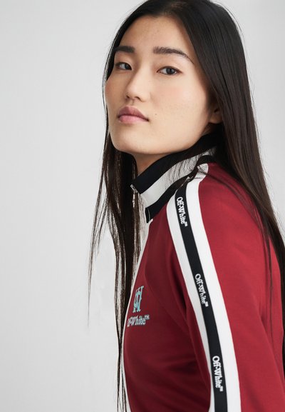 Maglia rossa con zip frontale e strisce laterali nere e bianche, con dettaglio logo. Tessuto liscio, collo alto, design aderente e maniche lunghe.