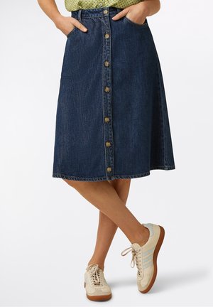 Person iført en midlang mørk denim knap-front nederdel med hænderne i lommerne og creme sneakers med blå striber, stående med krydsede ben.