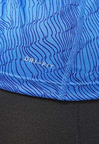 Sininen Dri-FIT-paita, jossa on aaltoileva kuvio tummemmassa sinisessä. Varustettu ommelluilla saumoilla ja sileällä pinnalla, yhdistettynä mustaan kangaspalaan alapuolella.