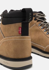Hnědé semišové kotníkové boty s černým textilním límcem, bílými prošívacími detaily, šedými tkaničkami a kontrastní béžovou gumovou podrážkou. Viditelná červená značka.