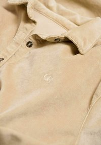 Nahaufnahme eines hellbeigen Hemdes mit einem geknöpften Kragen und einem gestickten Logo auf dem Stoff. Die Texturen sind weich und cordiert.