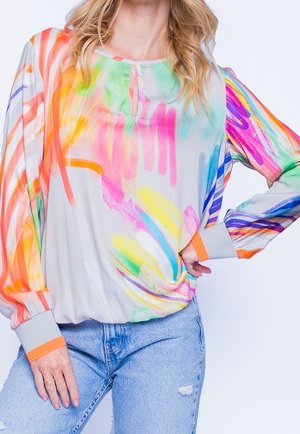 Femme portant un chemisier à manches longues avec des motifs abstraits colorés de coups de pinceau et un jean bleu clair déchiré, main sur la hanche.