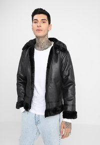 Chaqueta negra de cuero sintético con cuello y puños forrados de pelo sintético, cierre de cremallera y bolsillos laterales. Llevada sobre una camisa blanca y jeans de color azul claro.