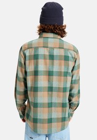 Camisa de franela a cuadros verde y marrón con cuello y botones. Mangas largas con puños redondeados, que muestran una textura tejida. Llevada con un gorro oscuro.