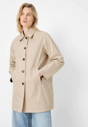 Frau mit blonden Haaren, die einen beigefarbenen, geknöpften Mantel mit Händen in den Taschen trägt, kombiniert mit hellblauen Jeans, schaut zur Seite.