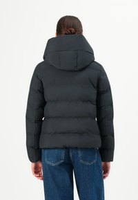 Femme aux cheveux bruns en chignon, portant une doudoune noire et un jean bleu, faisant face à un fond blanc.