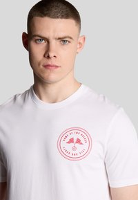 T-shirt bianco in cotone con un logo rotondo rosso, bandiere a scacchi e testo, design con colletto rotondo e maniche corte.