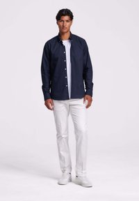 Man staat in een marineblauwe overhemd met knopen over een wit T-shirt, met witte broek en witte sneakers tegen een effen achtergrond.