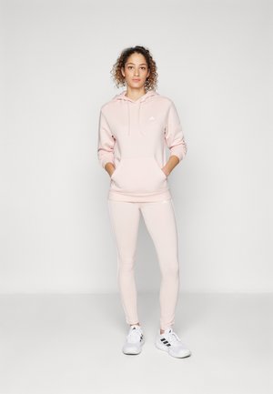 Sudadera de forro polar de color rosa claro con bolsillo frontal y cordón, combinada con leggings ajustados a juego y zapatillas deportivas blancas con detalles en negro.