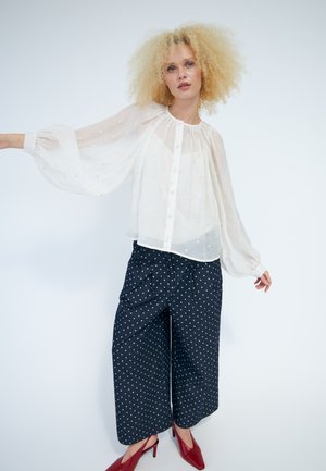 Femme aux cheveux blonds bouclés portant un chemisier blanc transparent, un pantalon large bleu marine à pois et des talons pointus rouges, posant devant un fond blanc.