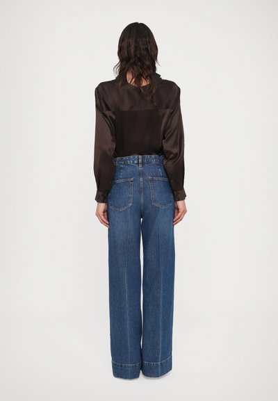 Femeie cu păr lung și întunecat, purtând o bluză din satin neagră introdusă în blugi evazați albaștri cu talie înaltă, stând cu fața spre spate pe un fundal alb.