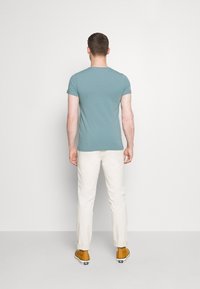 Tyrkysové tričko s krátkým rukávem z bavlny, kombinované s krémovými kalhotami a žlutými botami. Pohled zezadu, ukazující jednoduchý design.