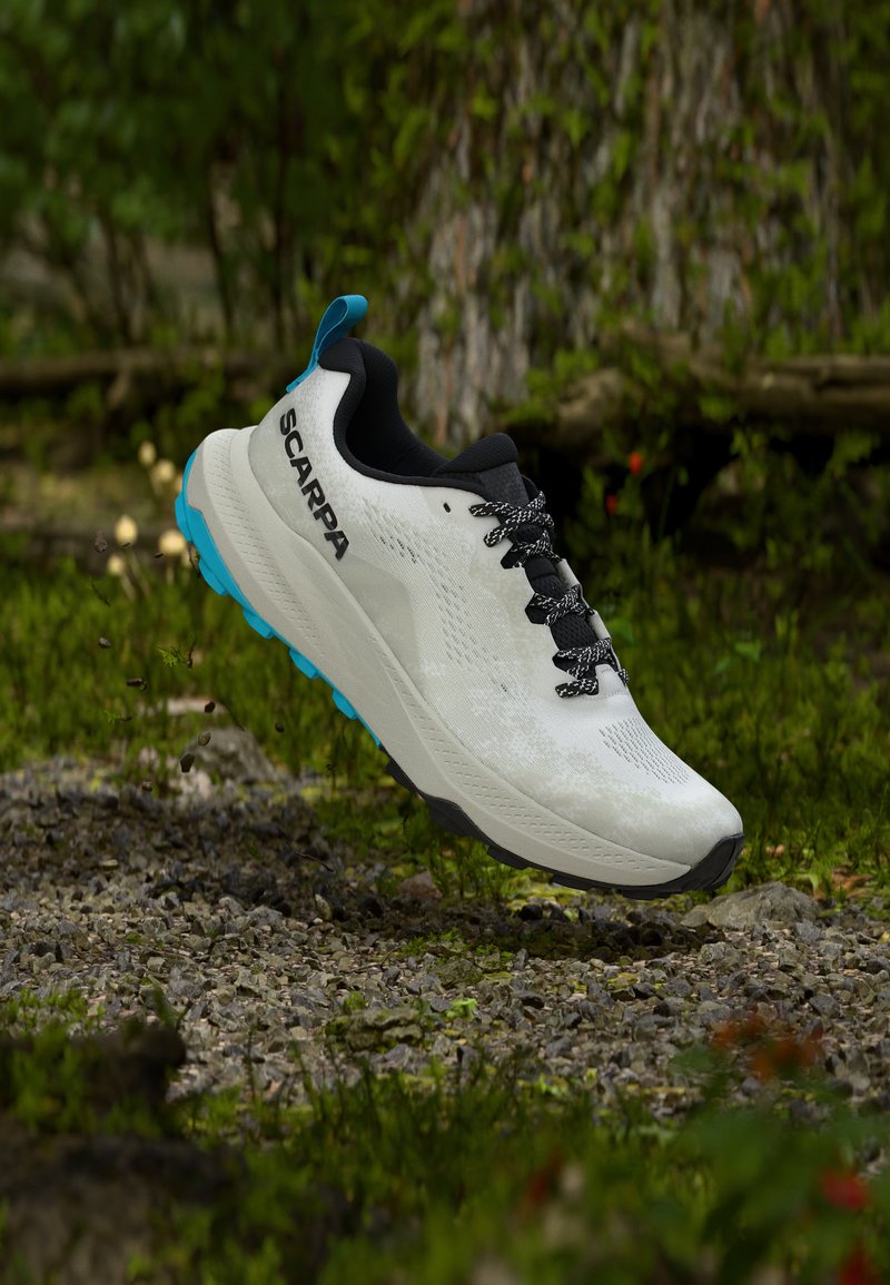 Λευκό και μπλε αθλητικό παπούτσι Scarpa με μαύρα κορδόνια να αιωρείται πάνω από ένα βραχώδες μονοπάτι στο δάσος.