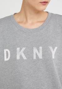 Osoba ve šedé mikině s bílými vyšitými písmeny "DKNY" viditelnými přes hruď, krk a dolní část obličeje je částečně vidět.