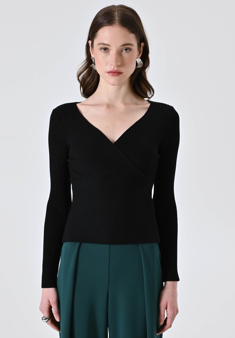 Maglietta nera a maniche lunghe con scollo a V, caratterizzata da una vita affusolata e una texture liscia, abbinata a pantaloni larghi verdi.