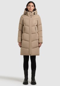 Bēšs pūkains mētelis ar kapuci, kas raksturots ar izšūtu dizainu, priekšējo rāvējslēdzēju, divām sānu kabatām un teksturētu apdari. Izskatu papildina melni zābaki.