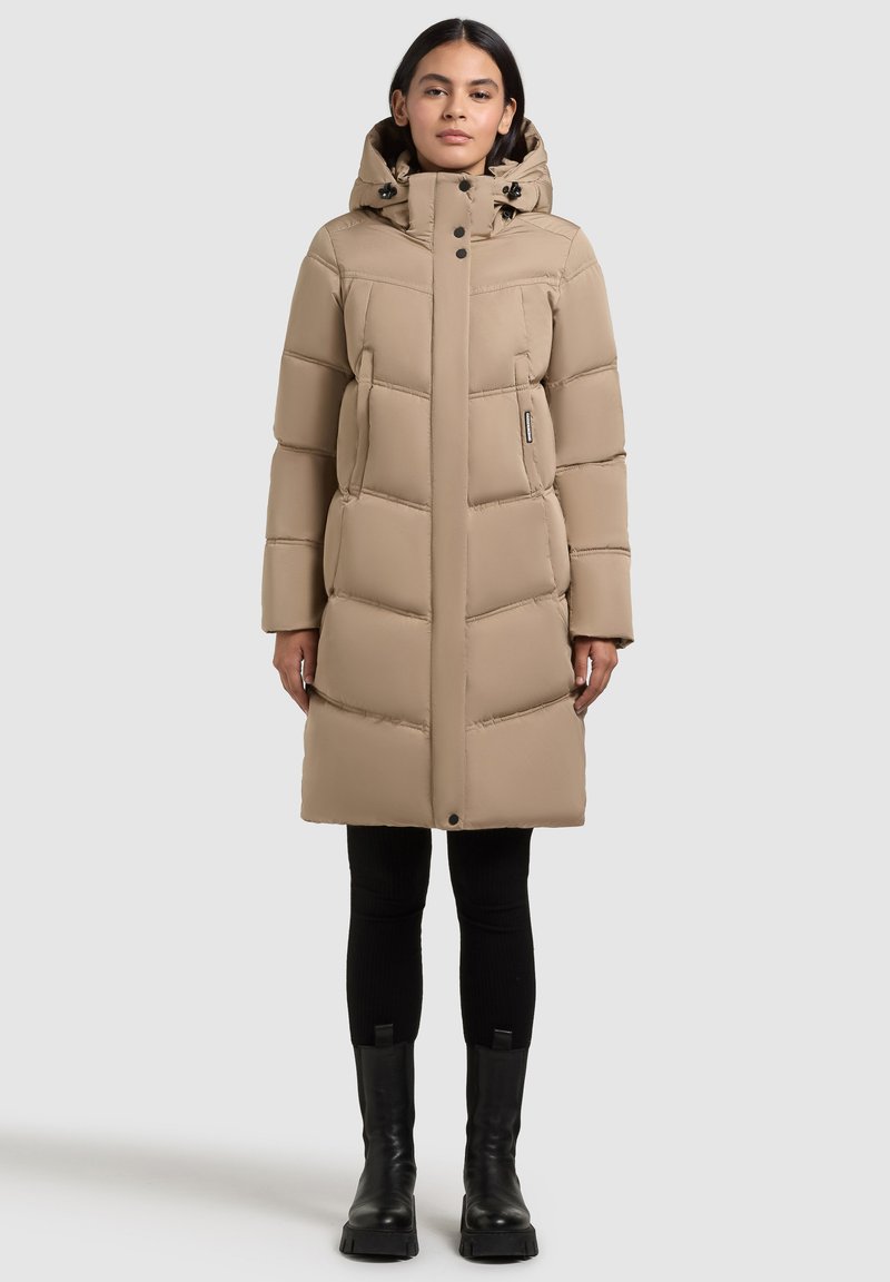 Bēšs pūkains mētelis ar kapuci, kas raksturots ar izšūtu dizainu, priekšējo rāvējslēdzēju, divām sānu kabatām un teksturētu apdari. Izskatu papildina melni zābaki.