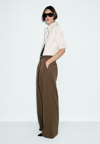Camicia oversize color beige chiaro, pantaloni larghi marroni e occhiali da sole neri. Tessuto morbido con una silhouette rilassata e tasche laterali.
