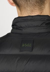 Svart puffjacka med hög krage, prydd med en grön präglad BOSS-logotyp på en fyrkantig svart lapp. Slät textur med kviltad design.