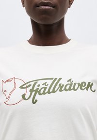 Κοντινό πλάνο ενός ατόμου που φοράει λευκό πουκάμισο με πράσινο λογότυπο «Fjällräven» και κόκκινο περίγραμμα κεφαλής αλεπούς στο στήθος.