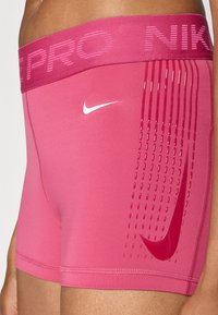 Calções de desporto cor-de-rosa com uma cintura plana que apresenta "NIKE PRO". Painel lateral texturizado com pequenas perfurações e um logótipo da Nike em branco.