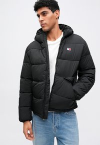 Jackett puf negru cu glugă, cu închidere prin fermoar, manșete elastice și un patch mic cu logo pe piept. Design călduros, capitonat.
