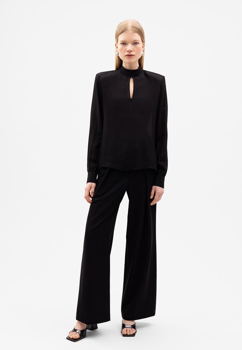 Schwarze Bluse mit hohem Kragen und Schlüssellochdetail, kombiniert mit lockeren schwarzen Hosen. Beide aus glattem Material mit minimalistischem Design.