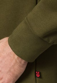 Olive green koszula z długim rękawem na mankiecie o prążkowanej teksturze, z małą czerwoną metką logo przy szwie. Szew jest starannie wykończony i trwały.