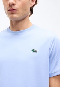 Άνδρας που φοράει ανοιχτό μπλε μπλουζάκι με μικρό πράσινο λογότυπο κροκόδειλου στο αριστερό στήθος, με λευκό φόντο.