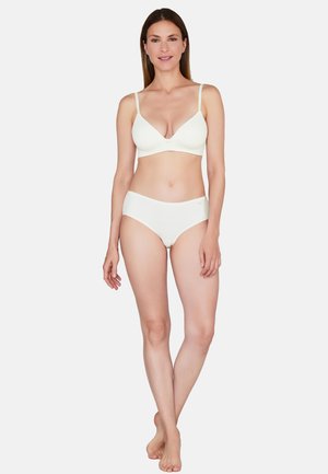 Woman standing barefoot wearing a white bra and matching white briefs, smiling with arms relaxed by her sides on a plain background.