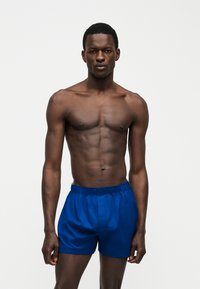 Homme à la peau foncée et en forme, debout devant un fond blanc, portant un short bleu à taille élastique, faisant face avec une expression neutre.