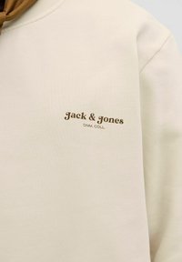 Beige színű pulóver, kerek nyakkivágással, puha textúrával és egy kis barna logóval, amelyen az áll: "Jack & Jones DNM. COLL." a mellkason.