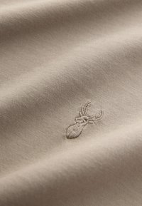 Tissu beige avec une petite tête de cerf brodée en détail avec un fil assorti montrant la texture et les plis.