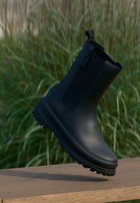 Botim de borracha preto até ao tornozelo com um acabamento texturizado, bico arredondado e sola robusta e pronunciada. Possui uma alça para fácil colocação.