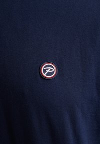 Námořnické tričko vyrobené z hladké látky, s kulatou vyšitou nášivkou s bílým a červeným obrysem a logem uprostřed.