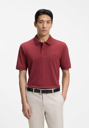 Hombre con una camisa polo color granate y pantalones beige claros con un cinturón negro, de pie frente a un fondo blanco liso.