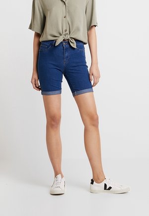 Persona che indossa pantaloncini di jeans blu scuro risvoltati, camicia a maniche corte verde oliva chiaro annodata e sneakers bianche con logo nero.
