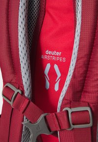 Röd ryggsäck med vadderad mesh, grå accenter och en spänne. Inkluderar "deuter AIRSTRIPES"-logotyp med en modern design.